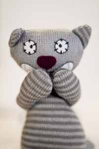 Doudou en laine grise aux pattes croisées sur fond épuré, invitant à la tendresse des cours de massage bébé lausanne à domicile ou au cabinet.